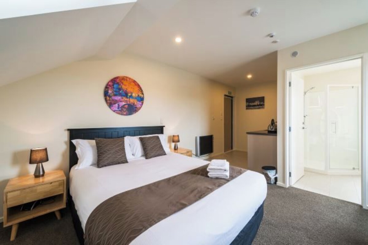 Habitación de hotel con una cama grande, dos mesitas de noche, una obra de arte colgada en la pared y una puerta que da al baño.