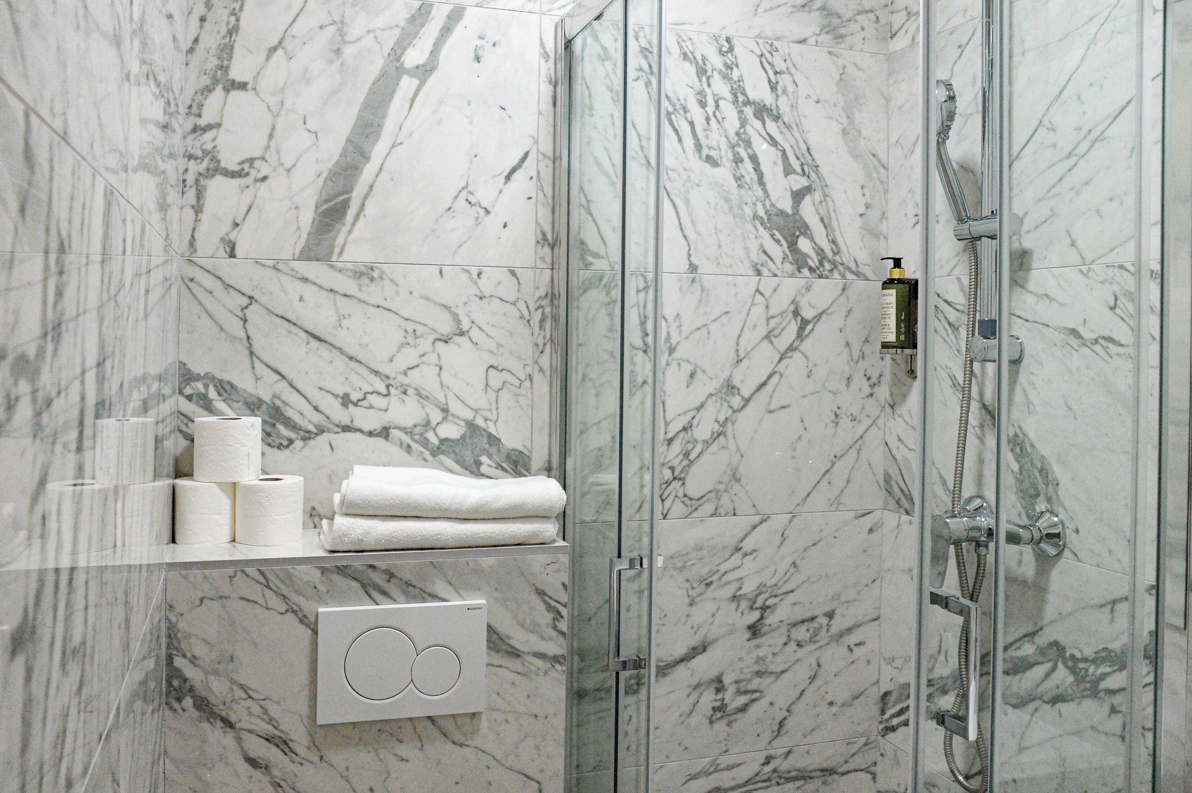 A bathroom with a glass shower enclosure, marble walls, a towel rack with towels, toilet paper, and a soap dispenser.