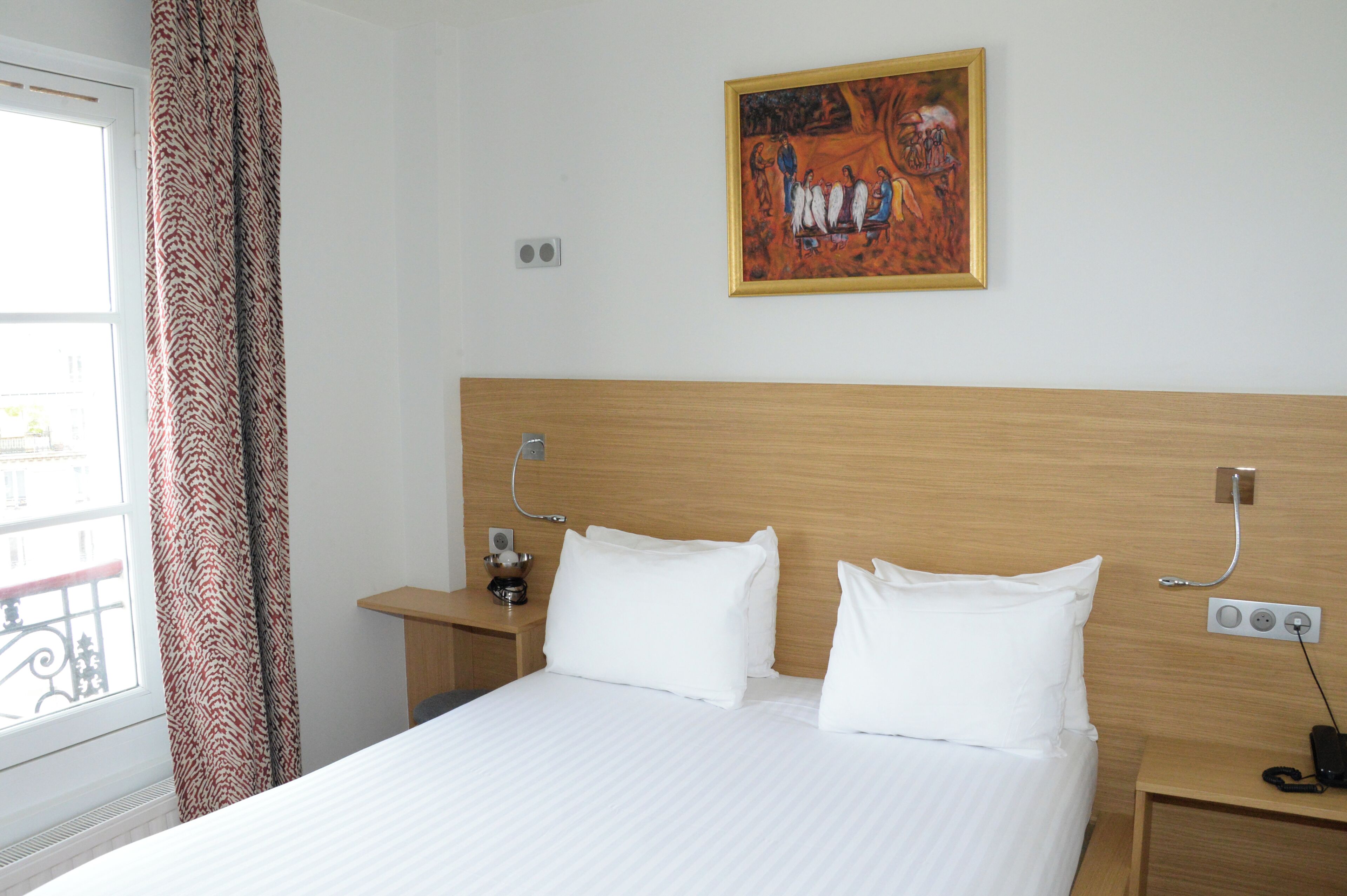 A hotel room with a bed, two pillows, a nightstand with a lamp, a painting on the wall, and a window with curtains.