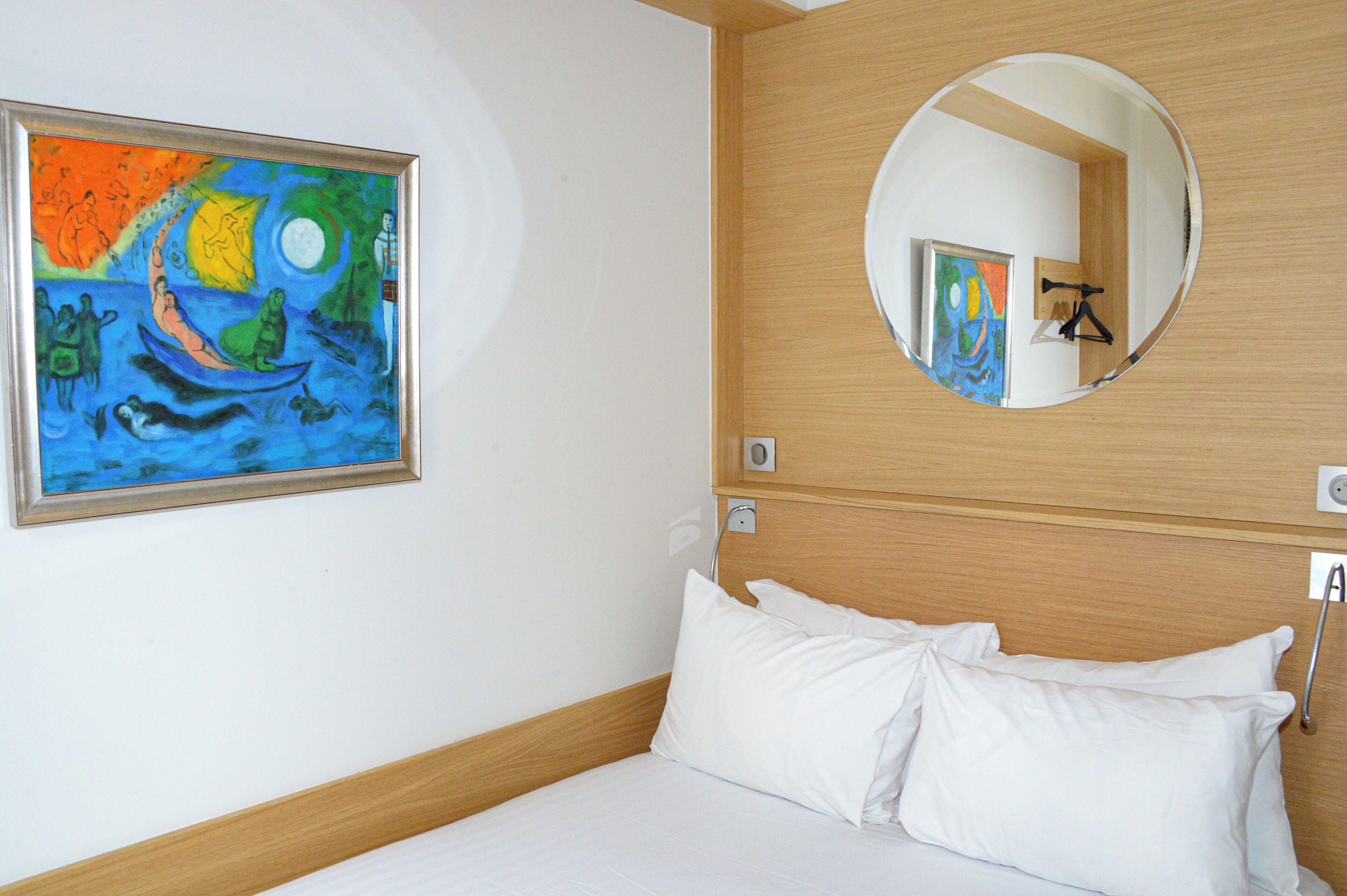 A hotel room with a bed, white pillows, a painting, a round mirror, and wooden paneling on the wall.