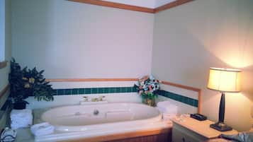 A hotel room with a large bathtub, a bedside table with a lamp, and a floral arrangement.