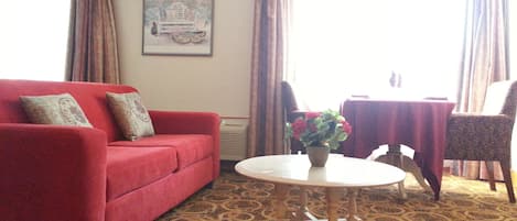 A living room with a red sofa, a round coffee table, a chair, and a table with a floral curtain.