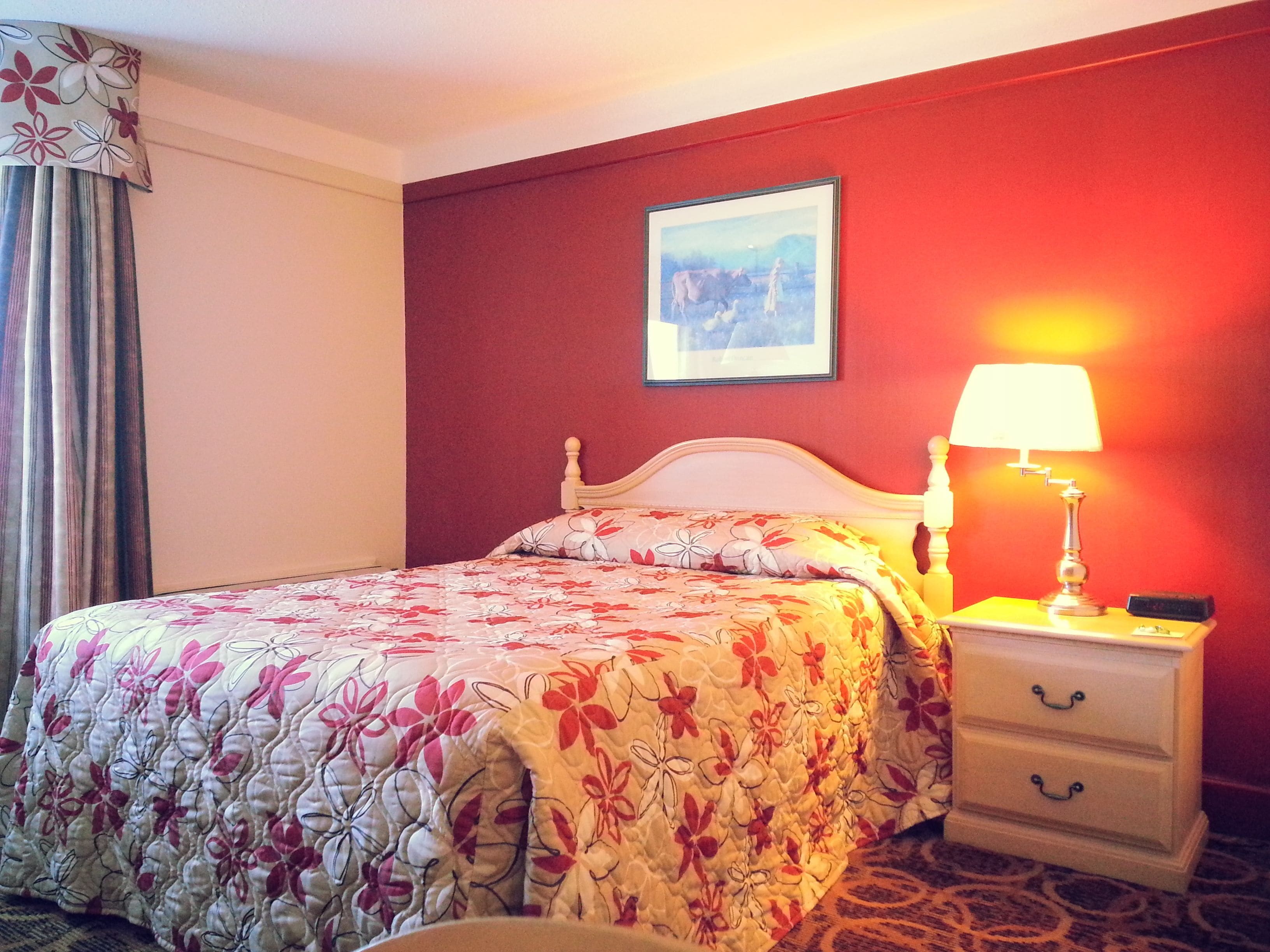 Une chambre d’hôtel avec un lit, une table de chevet avec une lampe, un tableau encadré au mur et une fenêtre avec des rideaux.
