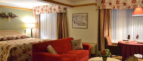 Una habitación de hotel con un sofá rojo, una mesa redonda con un jarrón de flores, una cama con ropa de cama floral y un cuadro en la pared.