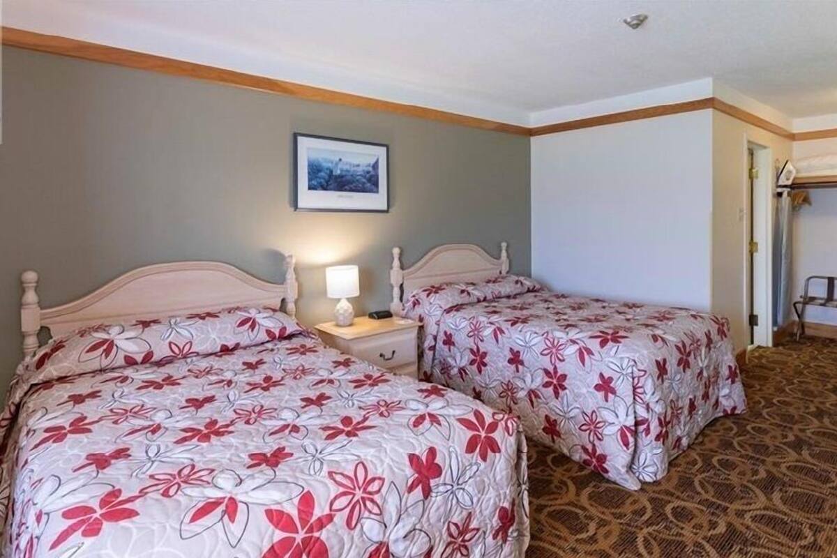 A hotel room with two beds, a nightstand with a lamp, a framed picture on the wall, and a bench near the door.