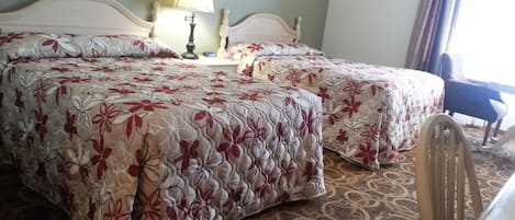A double bed with floral bedding, a bedside table with a lamp, and a chair in a room with patterned carpet and a window with curtains.