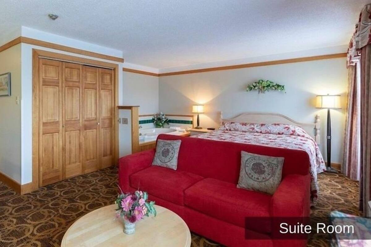 A suite room with a large bed, a red sofa, a wooden wardrobe, a bathroom with a bathtub, and a small round table with a vase of flowers.