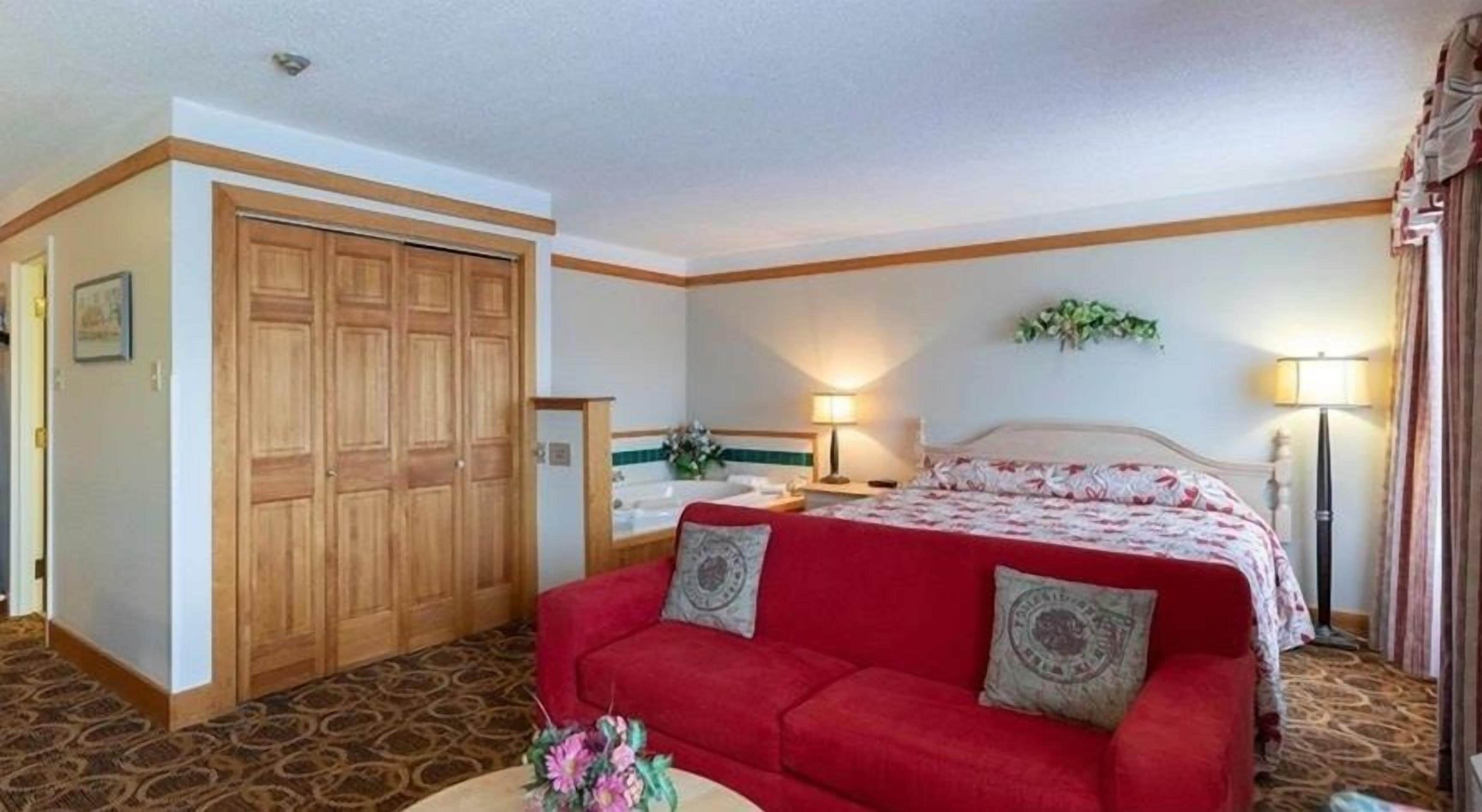 Une suite comprenant un grand lit, un canapé rouge, une armoire en bois, une salle de bain avec baignoire et une petite table ronde agrémentée d’un vase de fleurs.