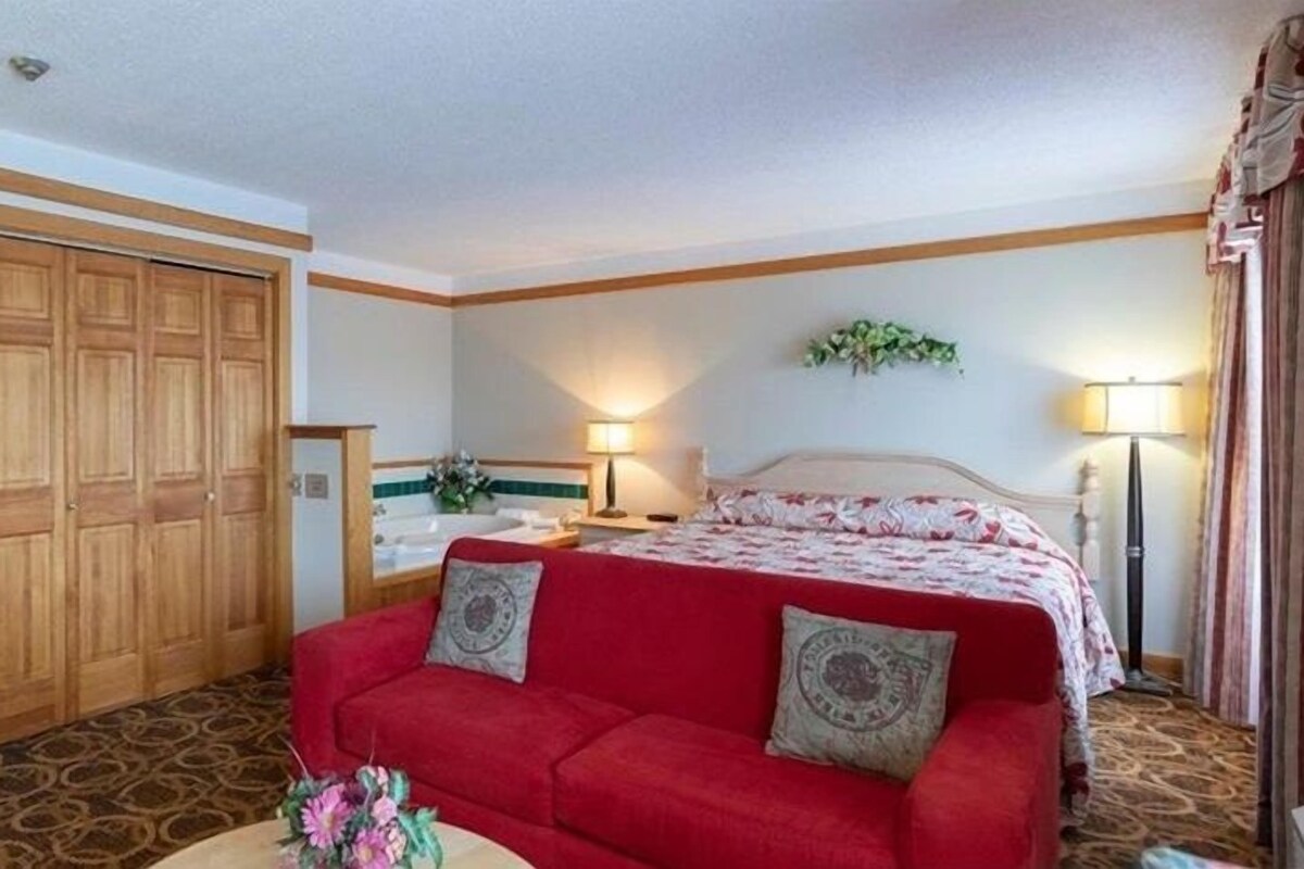Una suite con una cama grande, un sofá rojo, un armario de madera, un baño con bañera y una pequeña mesa redonda con un jarrón de flores.