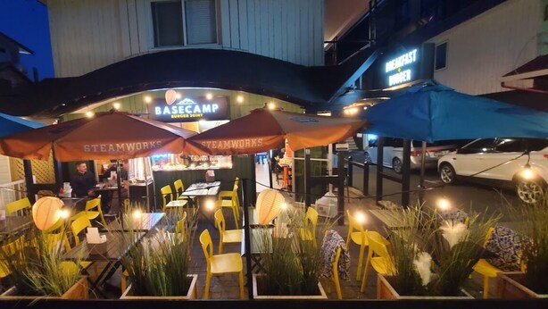 An outdoor dining area with yellow chairs and orange umbrellas, a sign for 'BASE CAMP BURGER 30INT', and a car parked nearby. - Harrison Spa Motel (Harrison Hot Springs)
