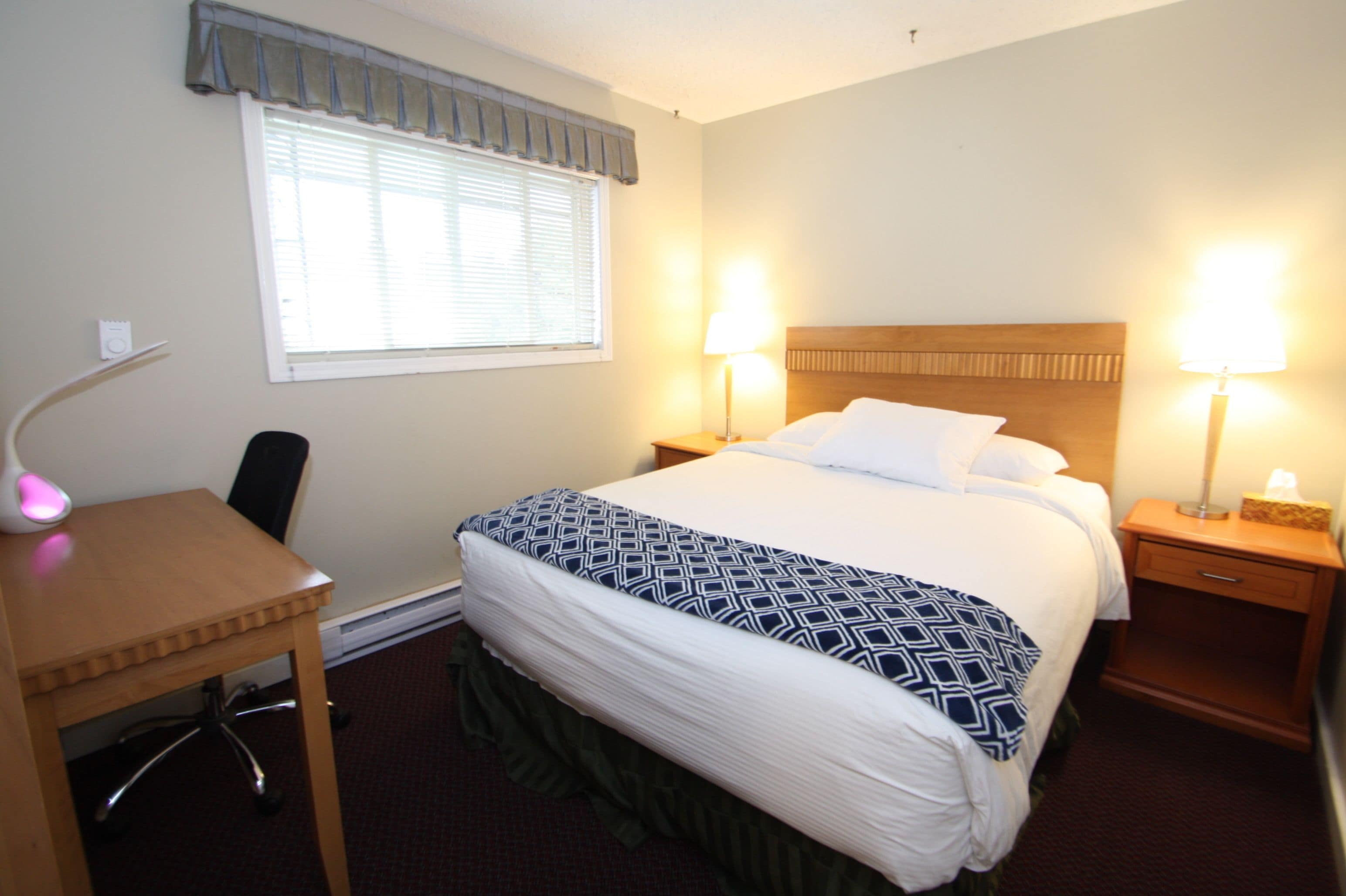 A hotel room with a bed, two bedside lamps, a desk with a chair, a window with blinds, and a wall-mounted telephone.