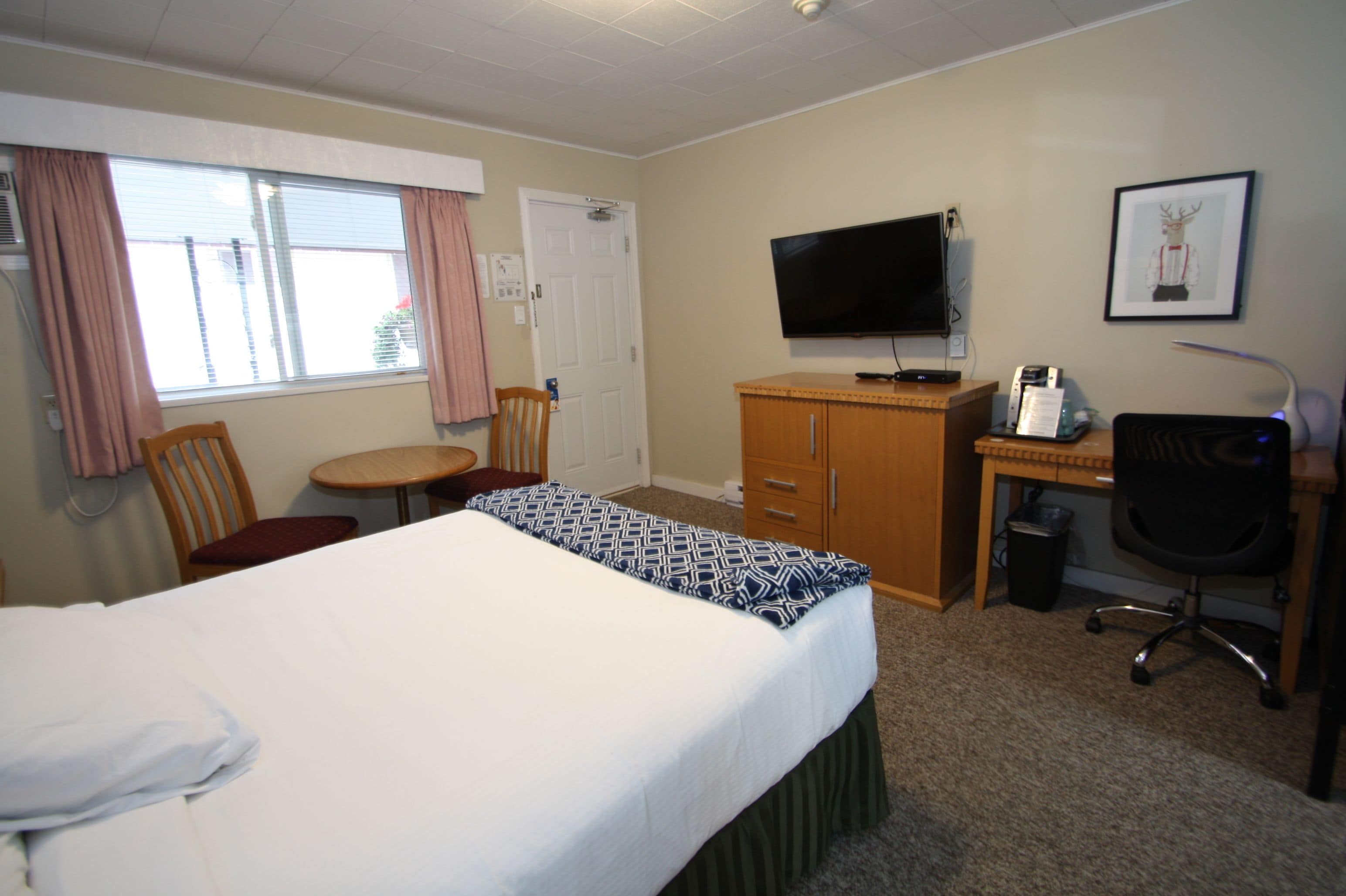 Quarto de hotel com cama, escrivaninha, cadeira, televisão e quadro na parede.