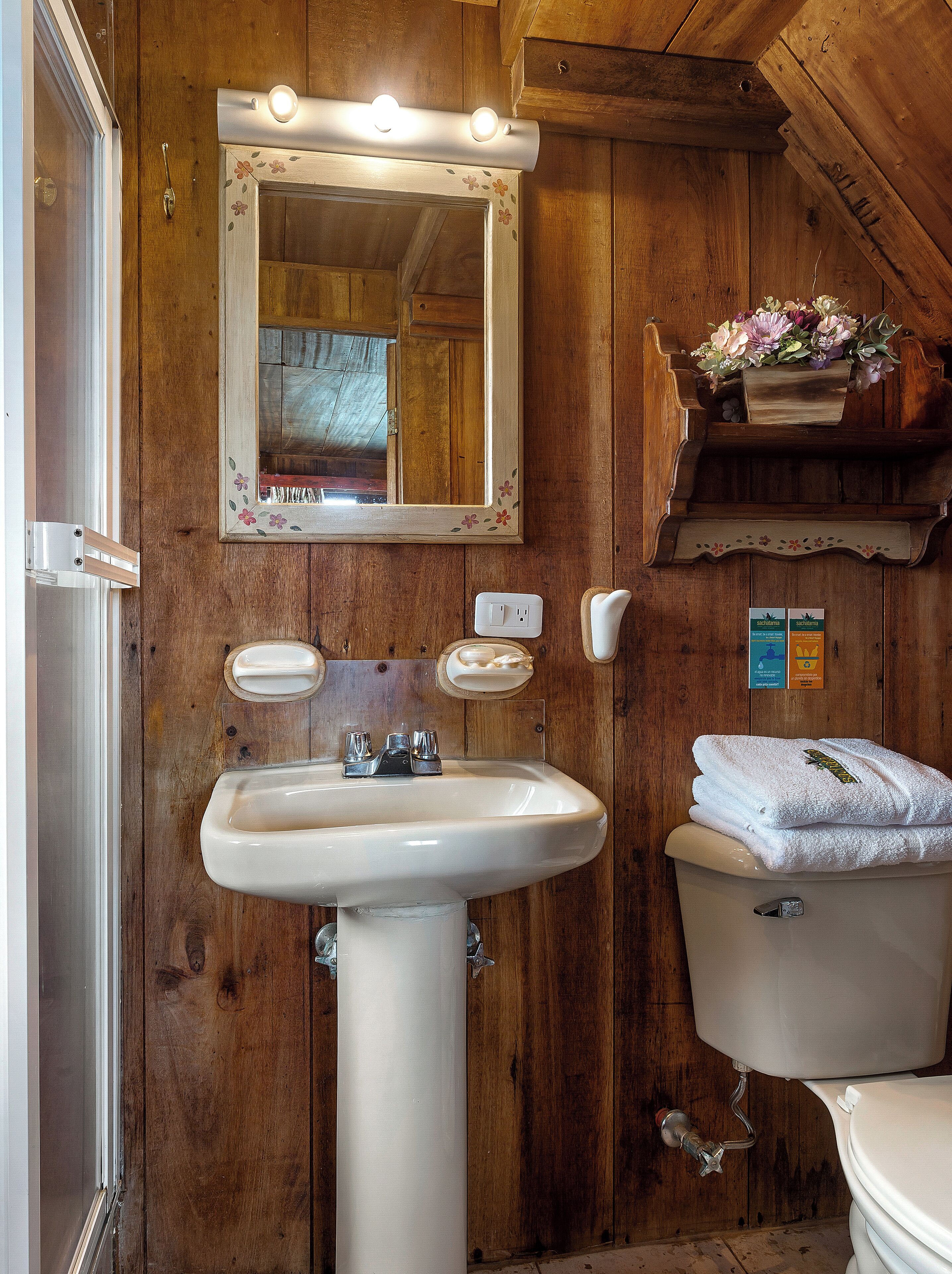 A bathroom with a wooden interior, a white sink, a mirror with lights, a toilet, and a shelf with towels and flowers.