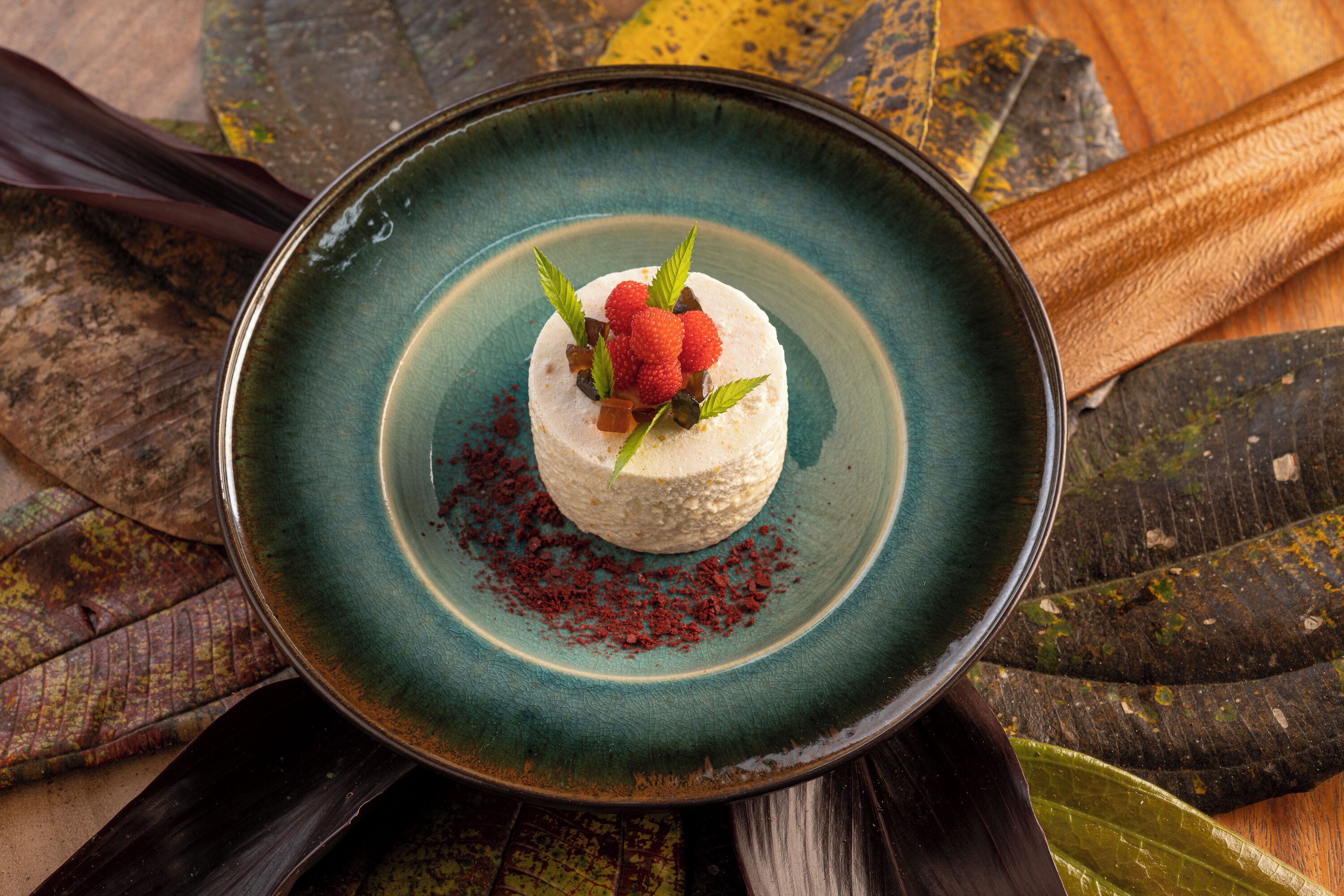 A dessert on a teal plate with scattered red powder, surrounded by autumn leaves.