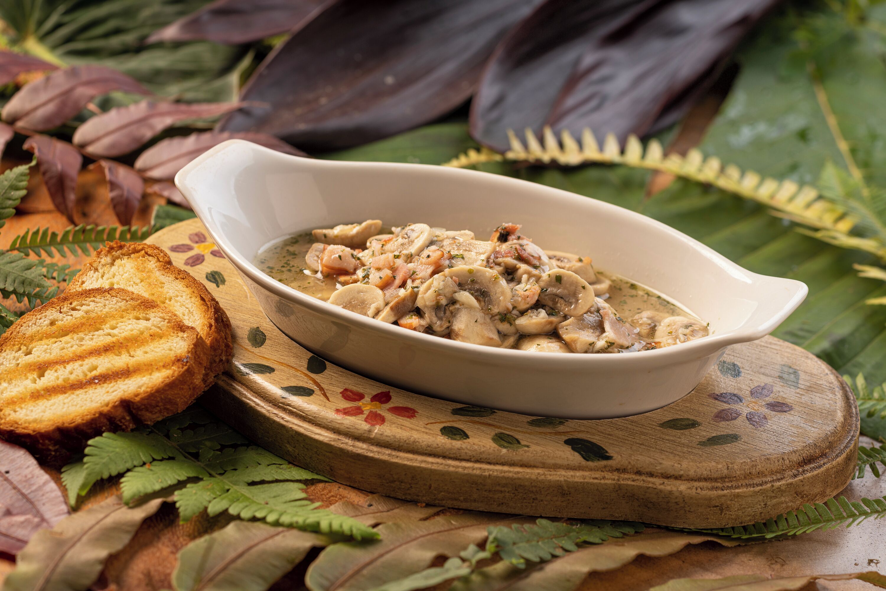 A bowl of mushroom soup on a wooden board with decorative leaves, accompanied by a slice of toasted bread.