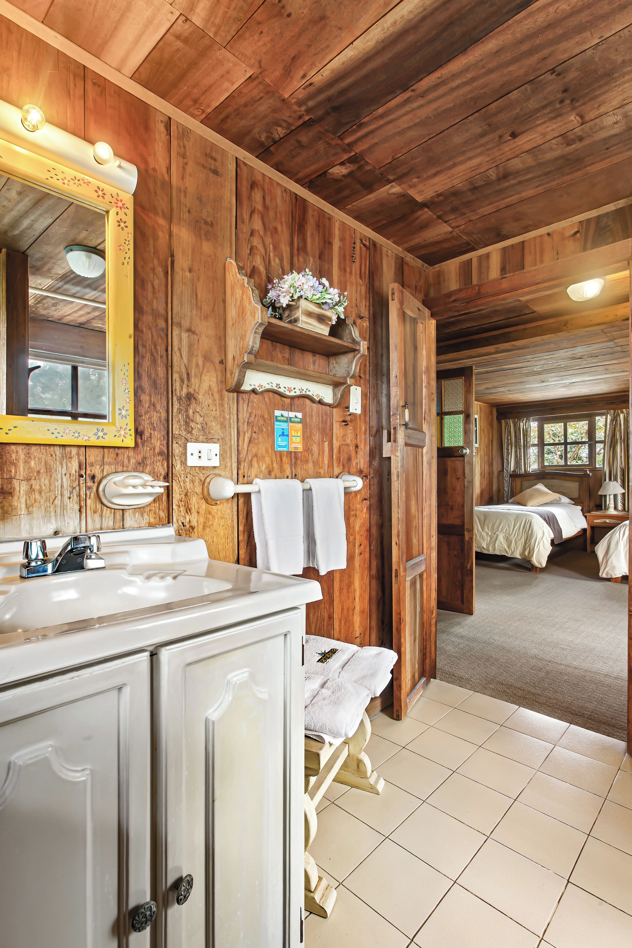 Interior de una cabaña de madera con baño, que incluye lavamanos, espejo y tocador, y un dormitorio con cama y ventana.