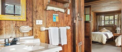 Interior de una cabaña de madera con baño, que incluye lavamanos, espejo y tocador, y un dormitorio con cama y ventana.