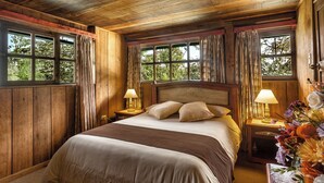 A wooden-paneled bedroom with a large bed, two bedside lamps, and a view of greenery through the windows. - Sachatamia Lodge (Mindo)