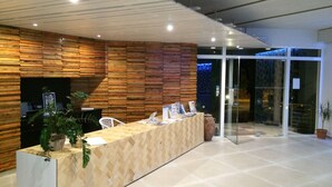 A reception desk with a wooden panel wall, a glass door entrance, and a potted plant. - Irina Beach Hotel (Kos)