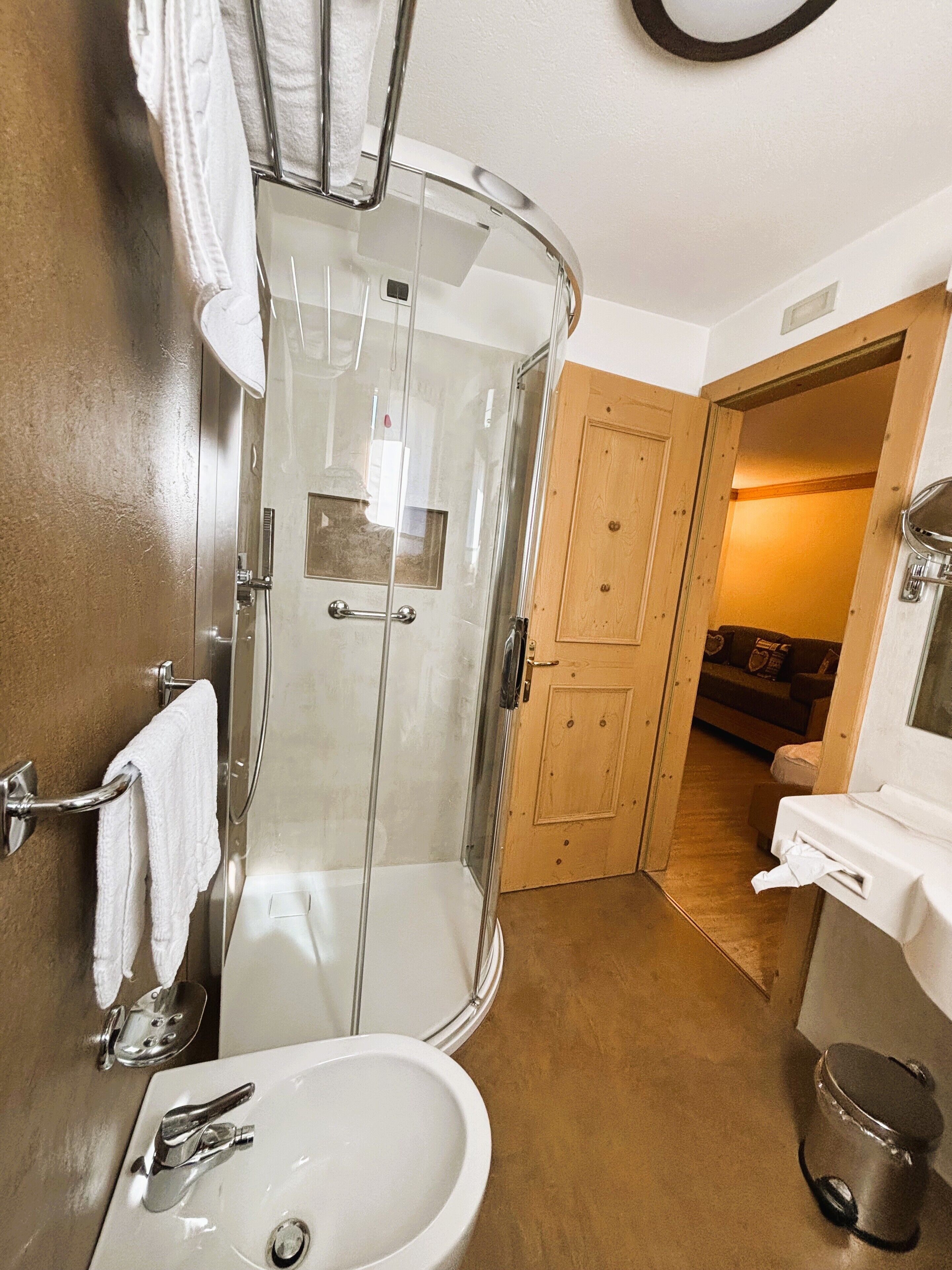 A bathroom with a glass-enclosed shower, a sink with a mirror, and a wooden door leading to a living area.