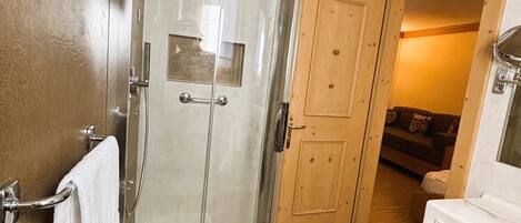 A bathroom with a glass-enclosed shower, a sink with a mirror, and a wooden door leading to a living area.