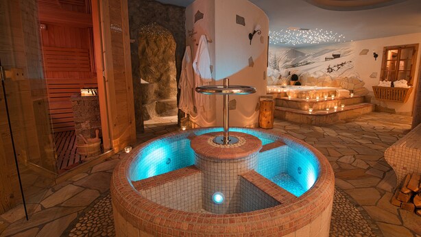 A spa area with a hot tub, wooden benches, and a stone wall with a mural. - Hotel Crosal (Livigno)