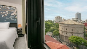 Hotellihuone, jossa on sänky, yöpöytä, lamppu ja näkymä kaupunkimaisemaan, johon kuuluu rakennuksia ja puita.