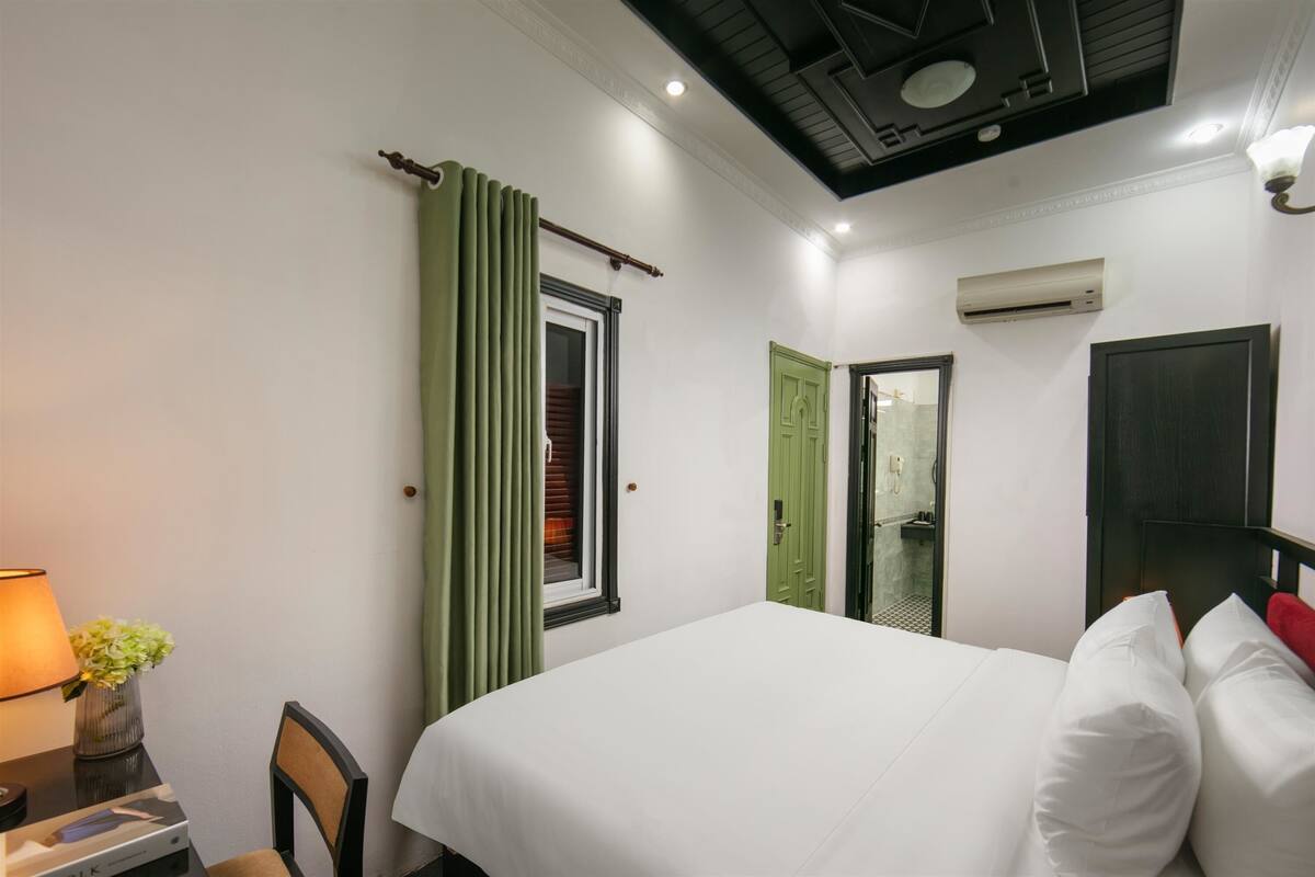 A hotel room with a bed, a desk with a lamp, a chair, a window with green curtains, and an air conditioning unit.