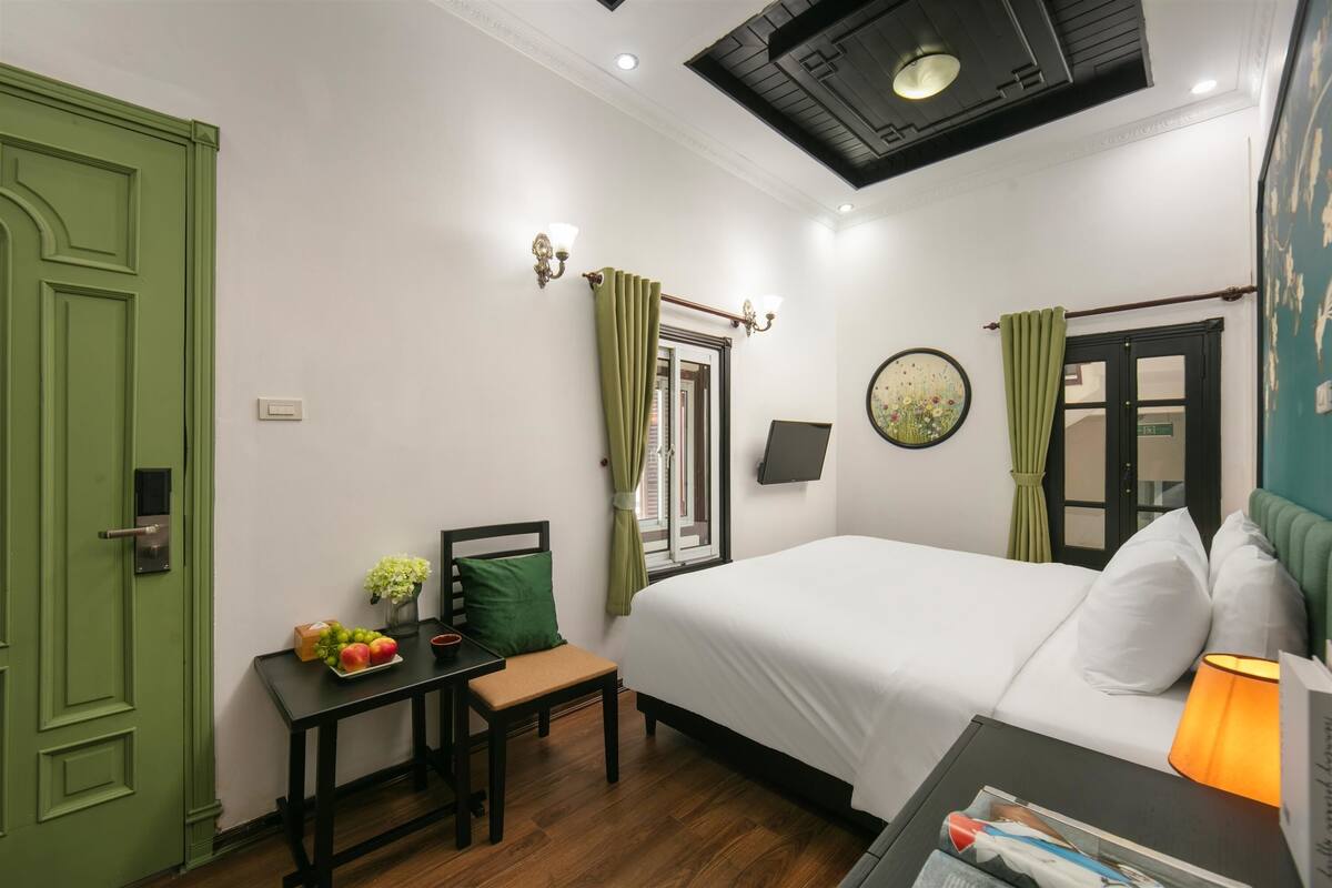 A hotel room with a bed, a desk with a chair, a small table with fruits, a lamp, and a green door.