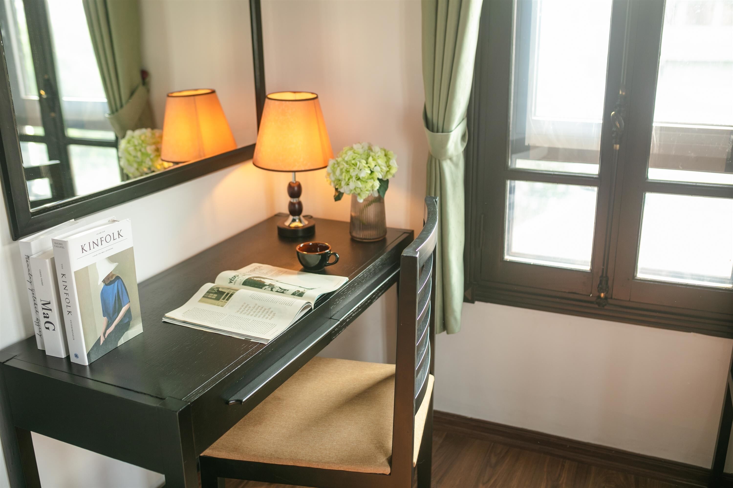 Una habitación de hotel con un escritorio de madera, un espejo, una lámpara, un jarrón con flores, una taza y libros.