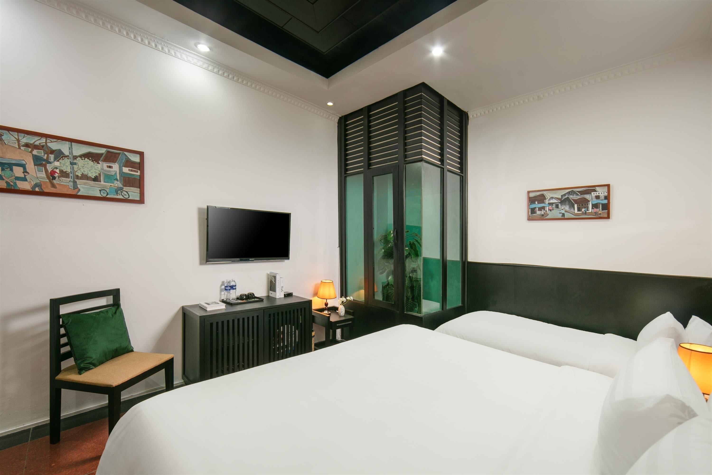 A hotel room with a bed, a desk with a lamp, a chair, a TV, and artwork on the walls.