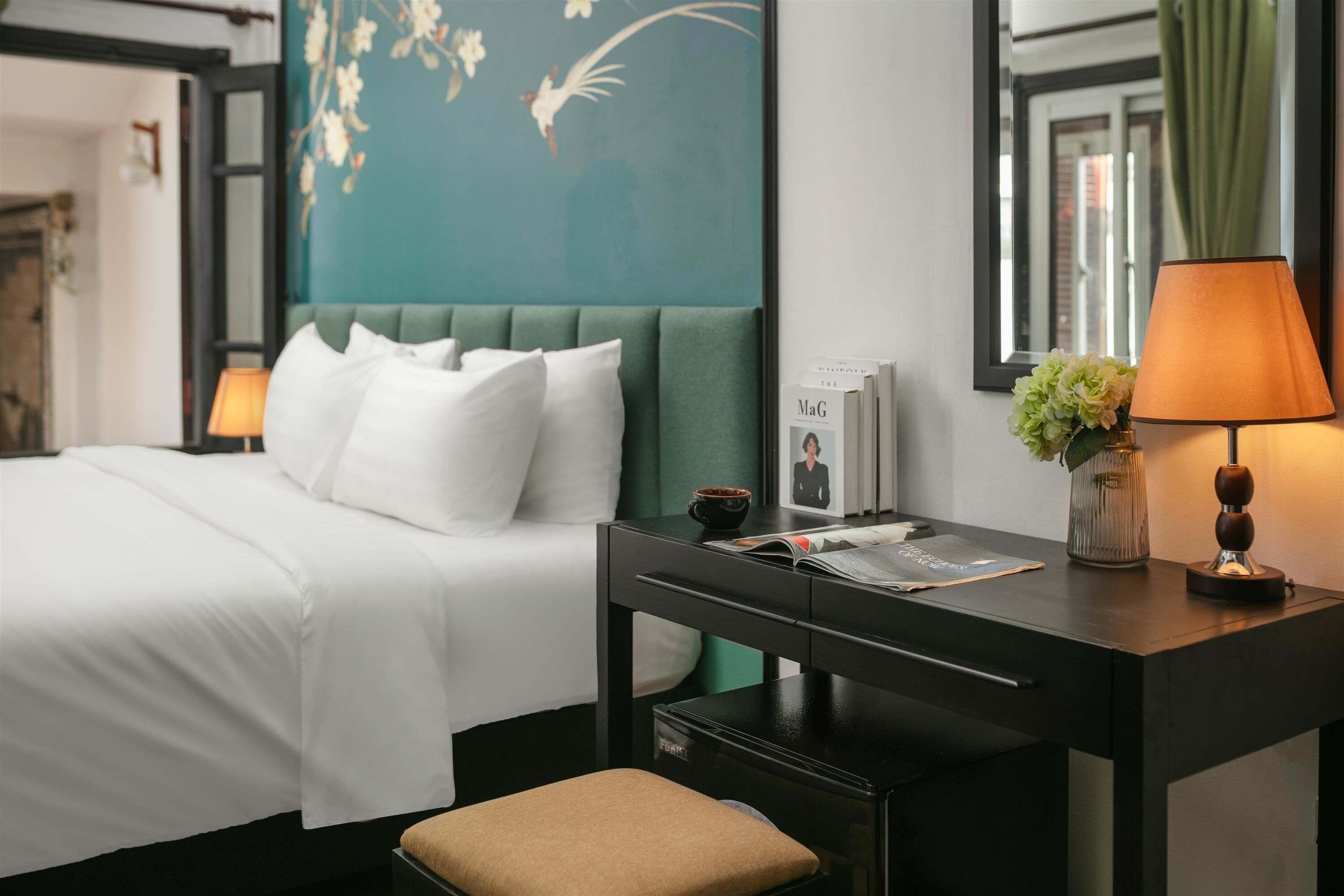 A hotel room with a bed, a desk, a lamp, a vase with flowers, and a framed photo on the desk.