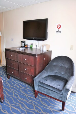 Hotelkamer met een donker houten dressoir, een flatscreen televisie, een grijs gestoffeerde fauteuil en een blauw kamerbreed tapijt.