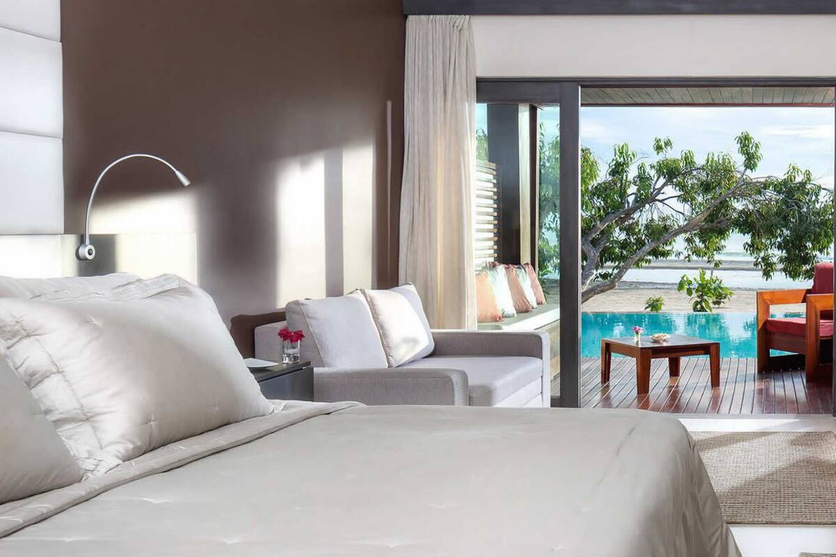 A modern hotel room with a large bed, a sofa, a small table, and a view of the beach and pool.