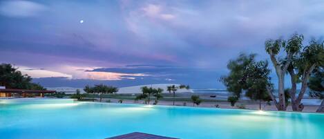 A swimming pool with a wooden deck, surrounded by palm trees and a view of the ocean at dusk.