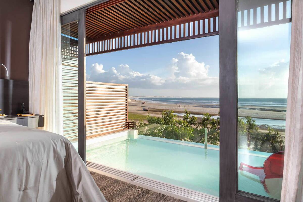 A bedroom with a large bed, a bedside table, and a view of a pool and beach.
