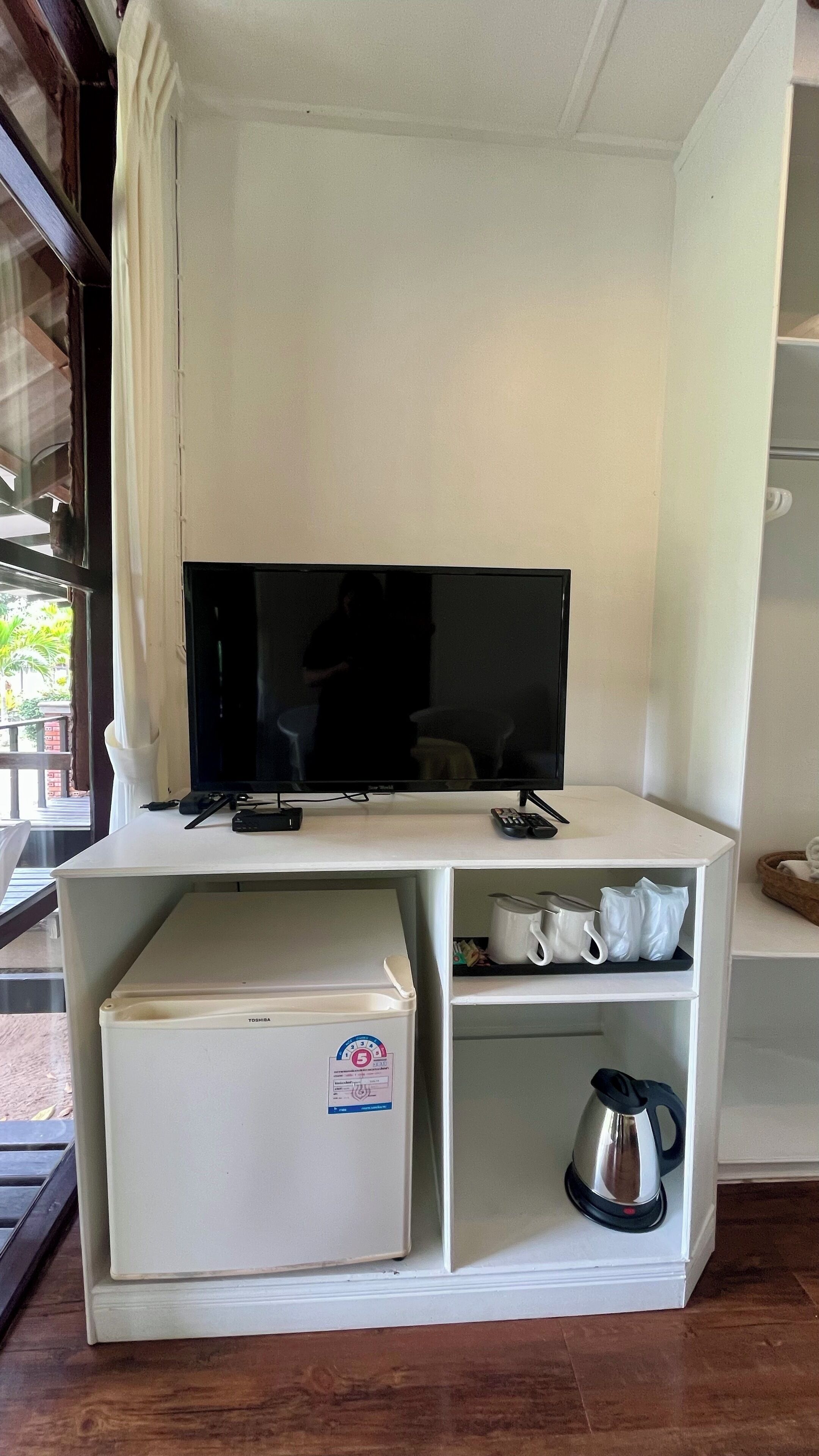 Un espacio habitable compacto con un televisor de pantalla plana sobre un mueble blanco, un refrigerador pequeño y una tetera.