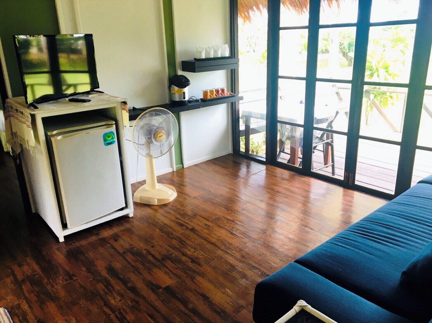 Una habitación con cama, un refrigerador pequeño, un televisor sobre un mueble bajo, un ventilador y una ventana con vistas al exterior.