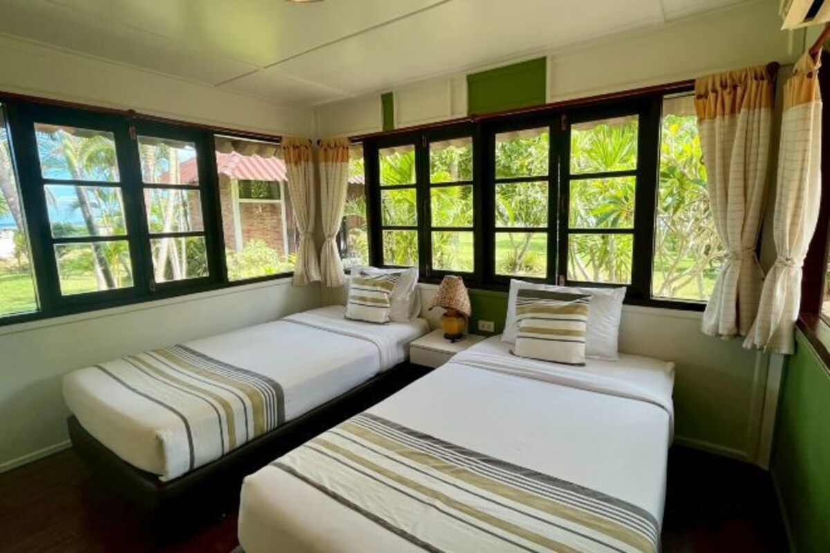 A room with two single beds, a nightstand with a lamp, and large windows with curtains.