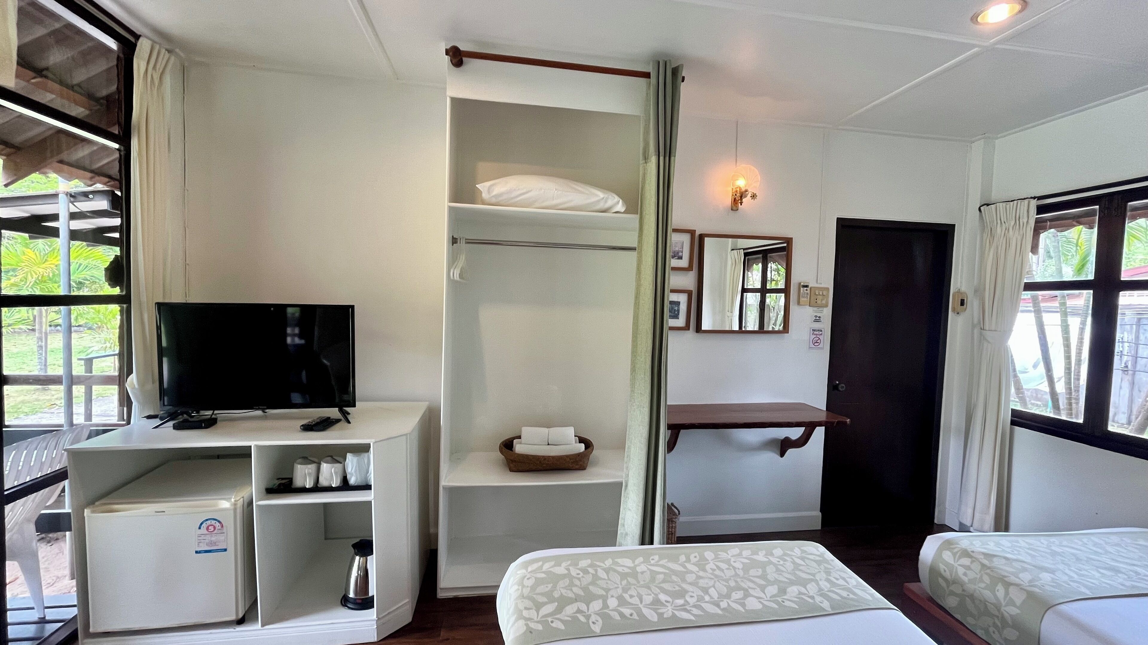 Hotelkamer met televisie, kleine koelkast, nachtkastje, kledingkast, raam met uitzicht op groen, en bed met bloemenprint beddegoed.