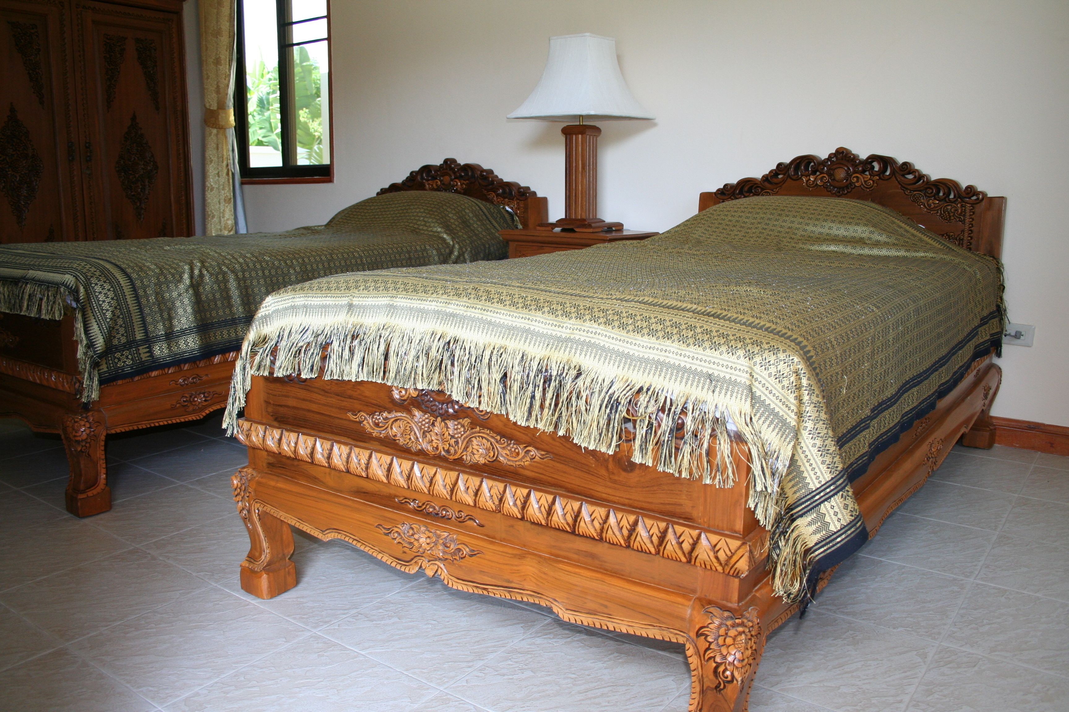 Duas camas de madeira com detalhes entalhados, uma mesa de cabeceira com abajur e uma janela com cortinas.