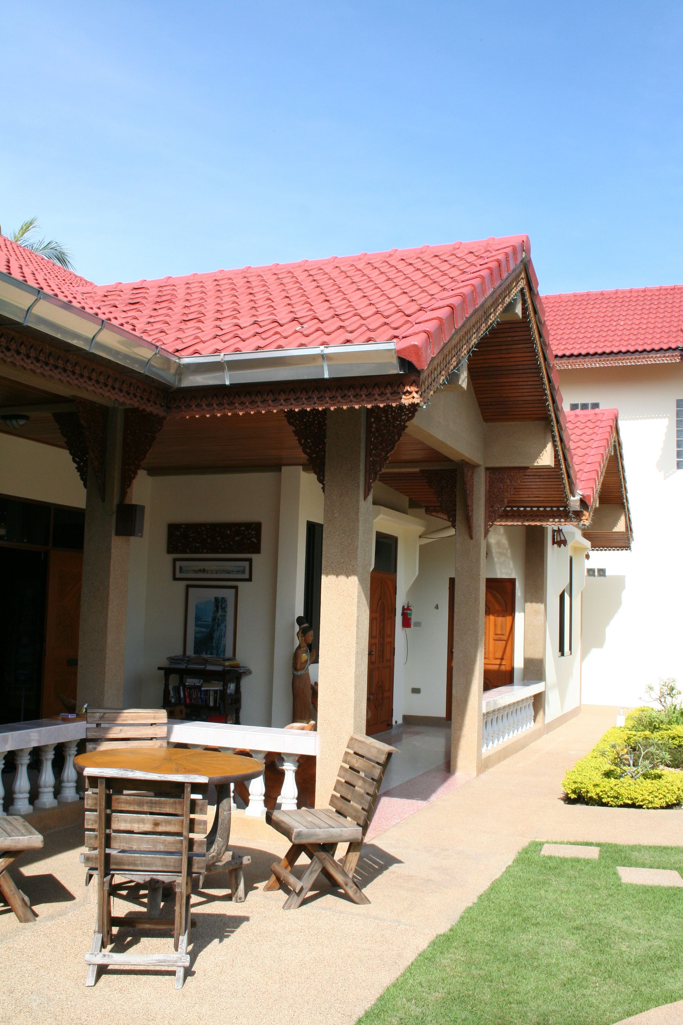 A covered outdoor seating area with a table and chairs, a small garden, and a building with a red-tiled roof.