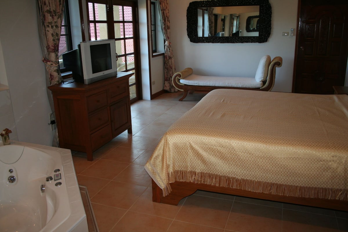 A bedroom with a bed, a TV on a wooden cabinet, a chair, a large mirror, and a bathtub.