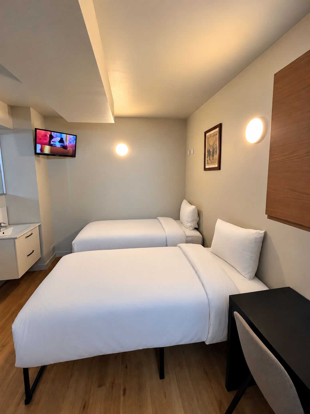 A hotel room with two beds, a desk, a TV, and a wall-mounted light.