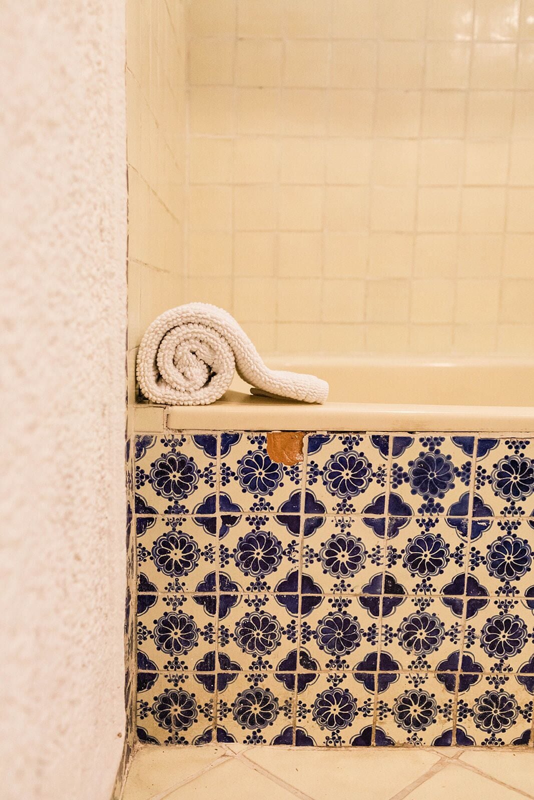 A bathroom with a white towel rolled up on a wooden ledge, a bathtub with a floral tile pattern, and a tiled floor.