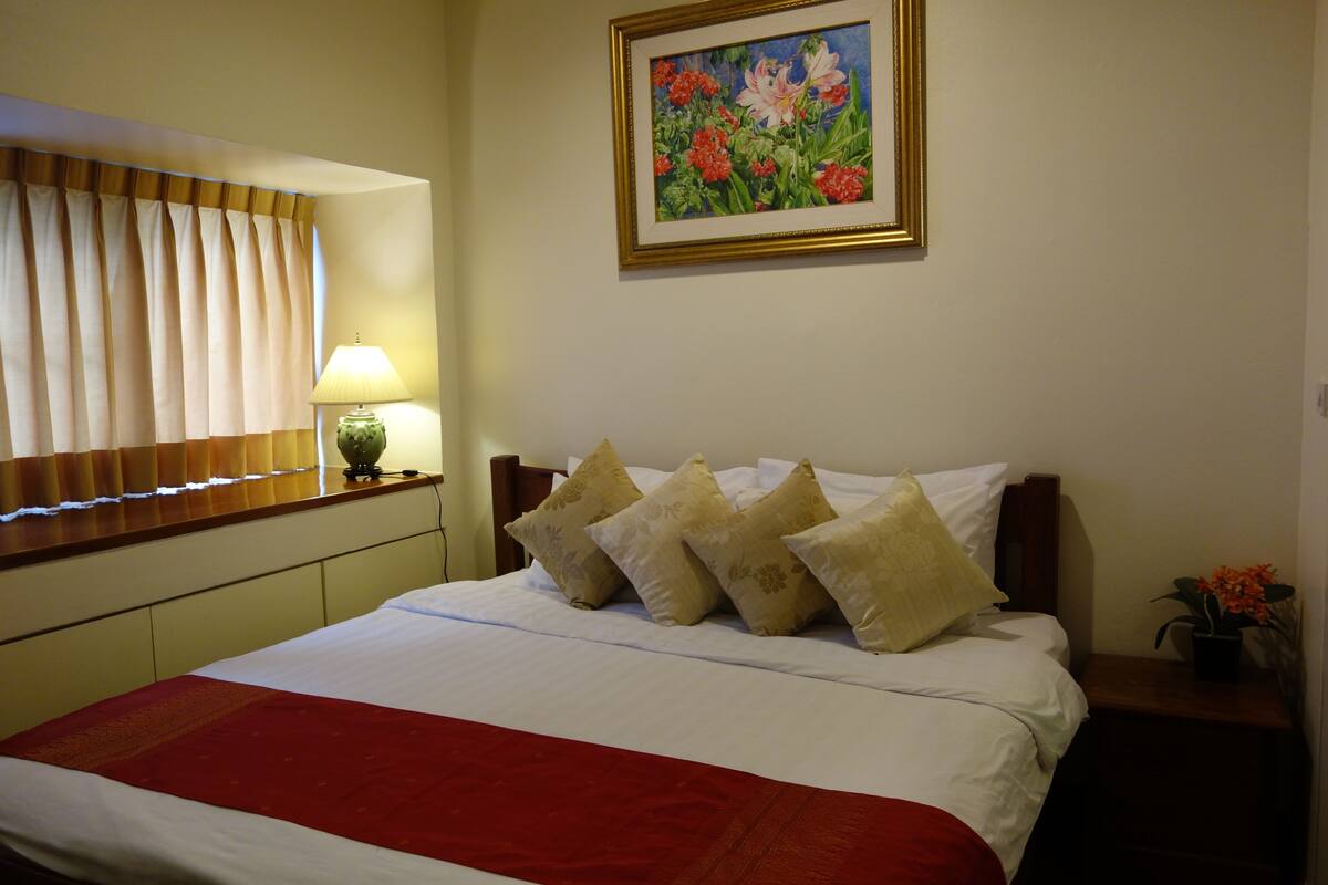 Un lit bien fait, avec une couverture rouge, une lampe de chevet et un tableau floral encadré accroché au mur.
