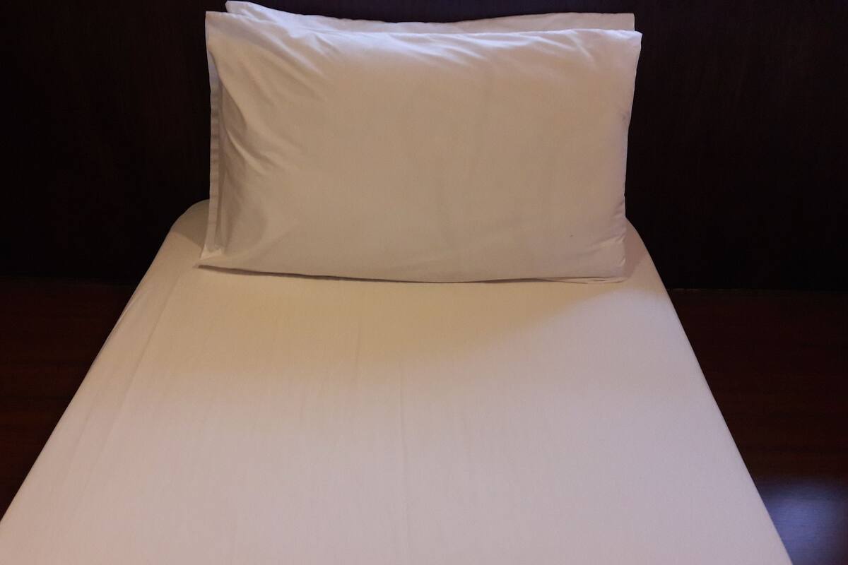 Un lit simple avec un matelas et un oreiller blancs, dans une pièce à la lumière tamisée et aux murs sombres.