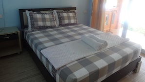 A neatly made bed with a checkered bedspread and pillows, a bedside table, and a view of the outdoors through a window. - Bird Bungalow (Ko Pha-ngan)