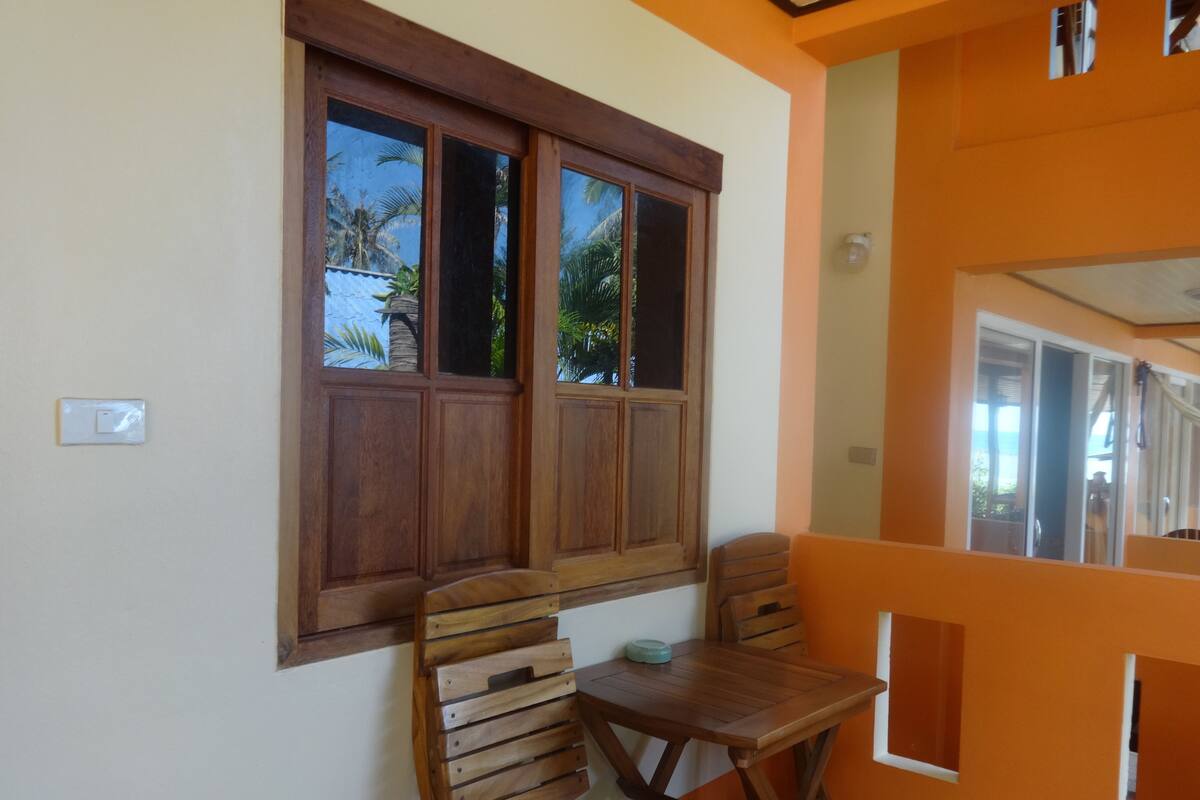 Una habitación con una mesa y sillas de madera, una ventana con marcos de madera y vistas a palmeras al exterior.