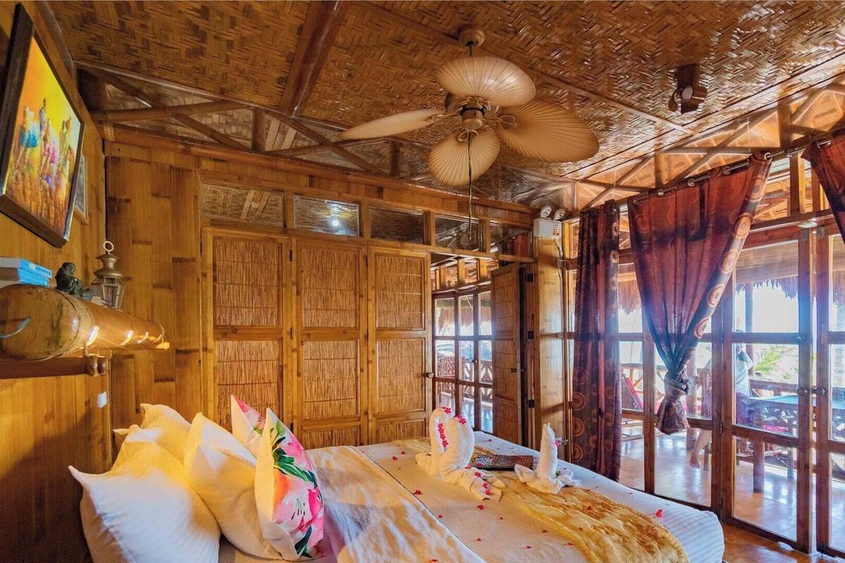 A room with wooden walls, a ceiling fan, a bed with floral pillows, and large windows with curtains.